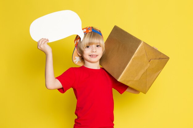 Une vue de face petit garçon en t-shirt rouge casquette colorée et un jean gris tenant la boîte sur le fond jaune