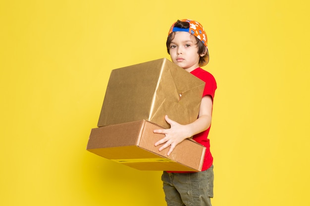 Une vue de face petit garçon en t-shirt rouge casquette colorée et pantalon kaki holding box sur le fond jaune