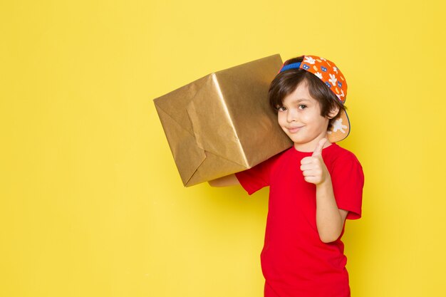 Une vue de face petit garçon en t-shirt rouge casquette colorée et pantalon kaki holding box sur le fond jaune