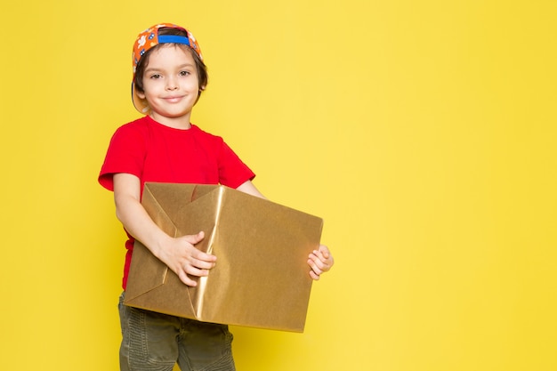 Une vue de face petit garçon en t-shirt rouge casquette colorée et pantalon kaki holding box sur le fond jaune