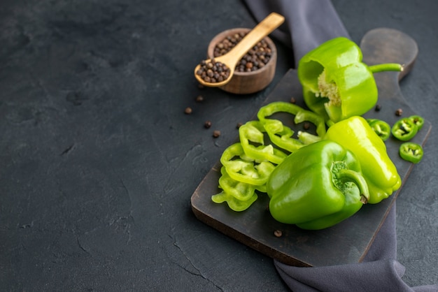 Photo gratuite vue de face de poivrons verts hachés entiers coupés sur une planche à découper en bois sur une serviette de couleur foncée sur une surface noire