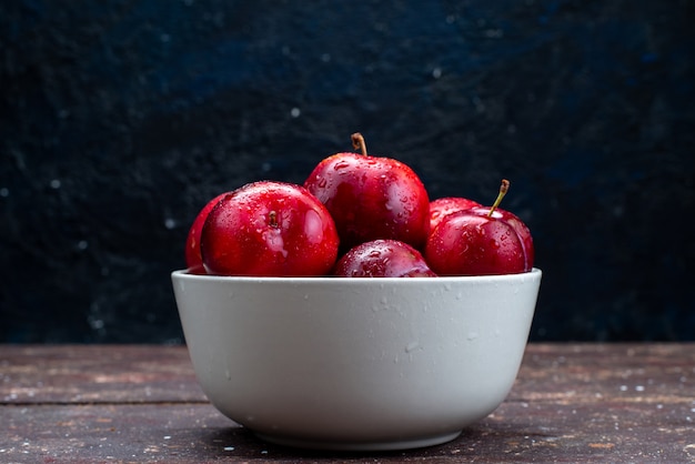 Une vue de face de prunes rouges fraîches moelleuses et mûres à l'intérieur de la plaque blanche sur le bureau en bois jus de pulpe de fruits