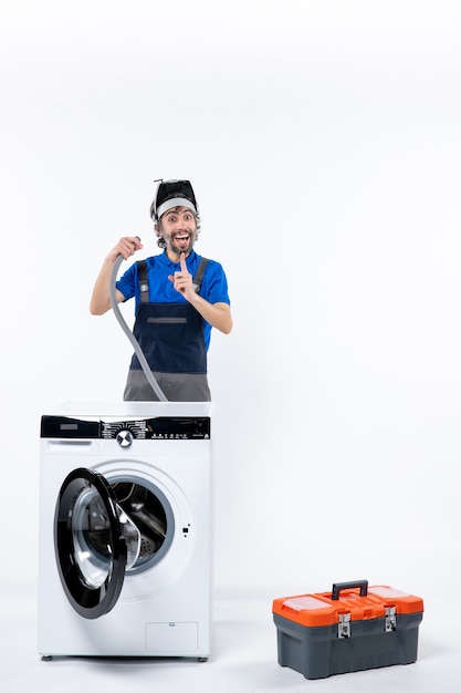 Vue de face d'un réparateur heureux en uniforme debout derrière une machine à laver tenant un tuyau sur un mur blanc