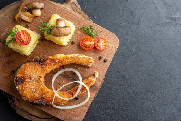 Vue de face d'un repas de poisson frit avec du fromage aux champignons et aux légumes sur une planche de bois sur le côté droit sur une surface noire en détresse