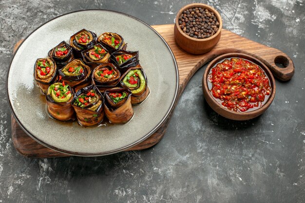 Vue de face rouleaux d'aubergines farcies dans une assiette ovale blanche poivre noir dans un bol sur une planche de service en bois avec poignée adjika sur fond gris