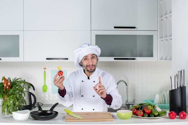 Vue de face satisfait chef masculin en uniforme brandissant la tomate dans la cuisine