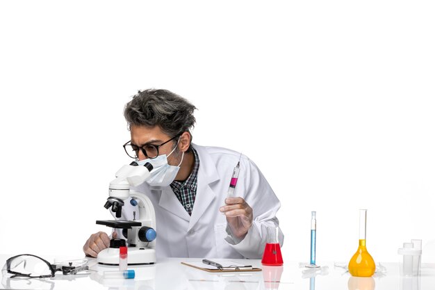 Vue de face scientifique d'âge moyen en costume médical blanc tenant l'injection prête et à l'aide d'un microscope