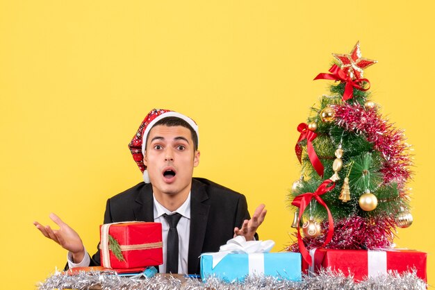 Vue de face se demandait bel homme avec bonnet de Noel assis à la table arbre de Noël et cadeaux