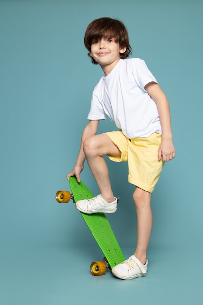 Photo gratuite une vue de face smiling cute kid riding skateboard vert en t-shirt blanc et short orange sur l'espace bleu