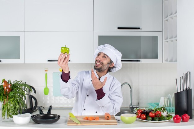 Vue de face souriant mâle cuisinier en uniforme brandissant du poivre vert dans la cuisine