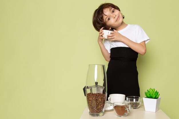 Une vue de face souriant mignon garçon en t-shirt blanc près de table avec du café sur le bureau de couleur pierre