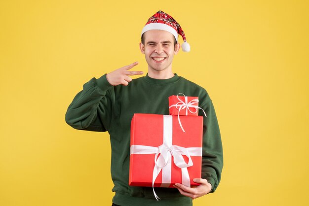 Vue de face sourit jeune homme avec cadeau de Noël faisant signe de la victoire debout sur jaune