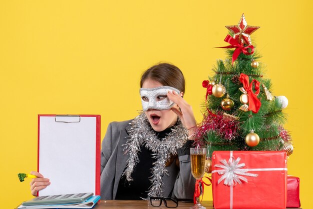 Vue de face surpris jeune fille avec masque de mascarade assis à la table tenant un arbre de Noël et des cadeaux