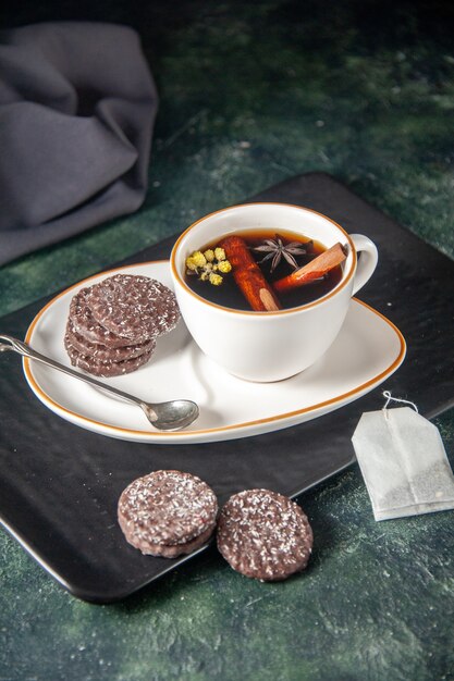 Vue de face tasse de thé avec des biscuits au chocolat sucré dans la plaque et le plateau sur la surface sombre verre cérémonie gâteau au sucre sucré couleur dessert