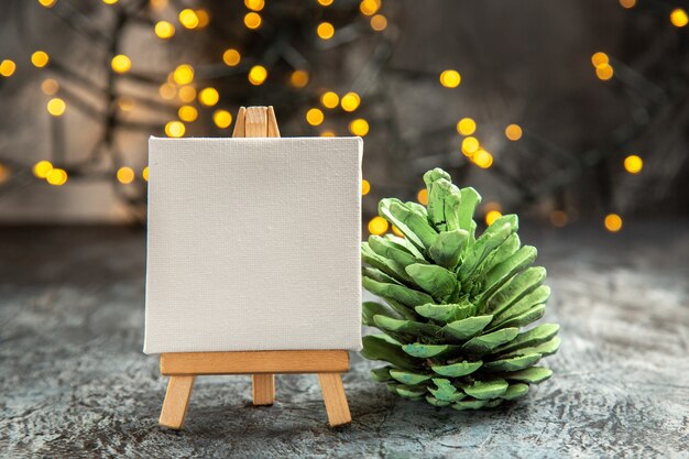 Photo gratuite vue de face toile blanche sur chevalet en bois lumières de noël pomme de pin verte sur fond sombre