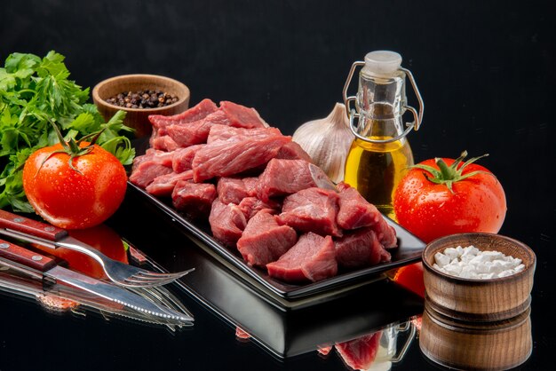 Vue de face des tranches de viande fraîche à l'intérieur d'un plateau noir avec des tomates et des légumes verts sur une table noire