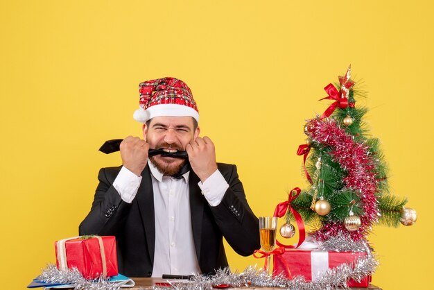 Vue de face travailleur masculin assis derrière sa table avec des cadeaux sur jaune