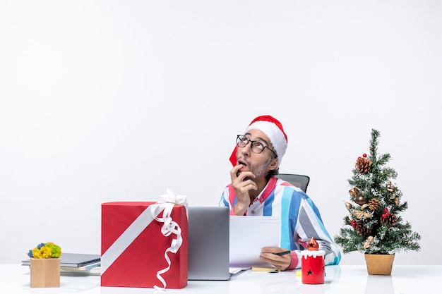 Vue de face travailleur masculin avec bonnet de noel assis à sa place avec un ordinateur portable et des fichiers contenant des documents