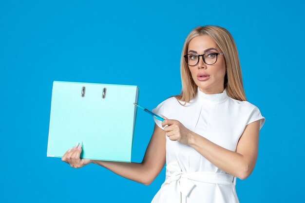 Vue de face d'une travailleuse en robe blanche tenant un dossier bleu sur un mur bleu