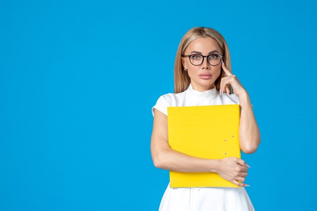 Vue de face d'une travailleuse en robe blanche tenant un dossier jaune sur un mur bleu