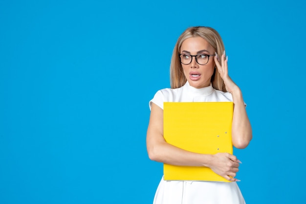 Vue de face d'une travailleuse en robe blanche tenant un dossier jaune sur un mur bleu