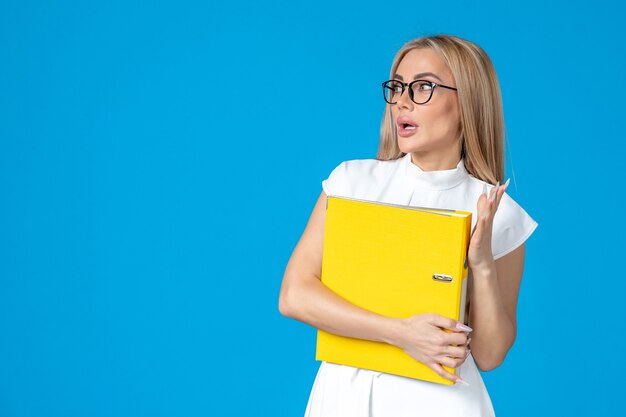 Vue de face d'une travailleuse en robe blanche tenant un dossier jaune sur un mur bleu