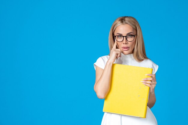 Vue de face d'une travailleuse en robe blanche tenant un dossier jaune sur un mur bleu