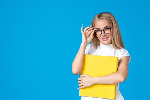 Vue de face d'une travailleuse en robe blanche tenant un dossier jaune sur un mur bleu