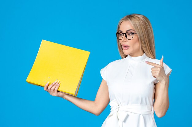 Vue de face d'une travailleuse en robe blanche tenant un dossier jaune sur un mur bleu