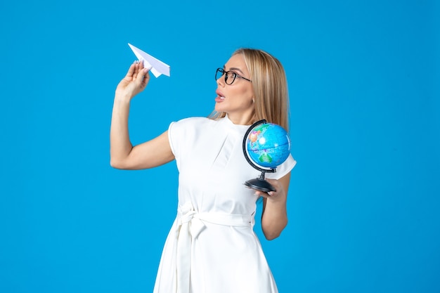 Vue de face d'une travailleuse en robe blanche tenant un globe terrestre et un avion en papier sur un mur bleu