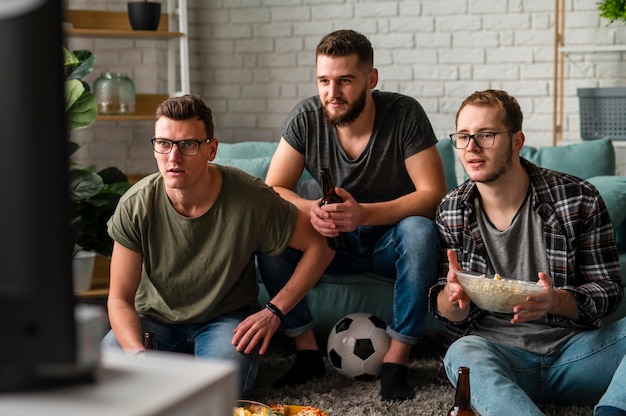 Vue de face de trois amis masculins à regarder le sport à la télévision ensemble tout en ayant de la bière et des collations
