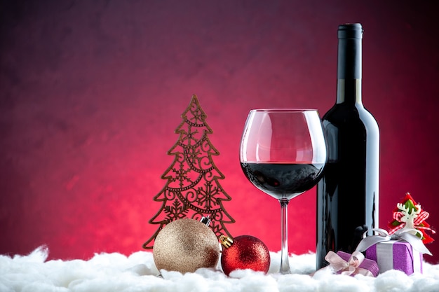 Photo gratuite vue de face un verre de vin bouteille de vin jouets de boule de noël sur fond rouge foncé