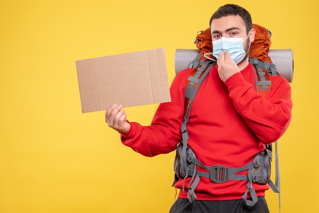 Vue de face d'un voyageur confiant portant un masque médical avec sac à dos pointant une feuille sans écrire sur fond jaune