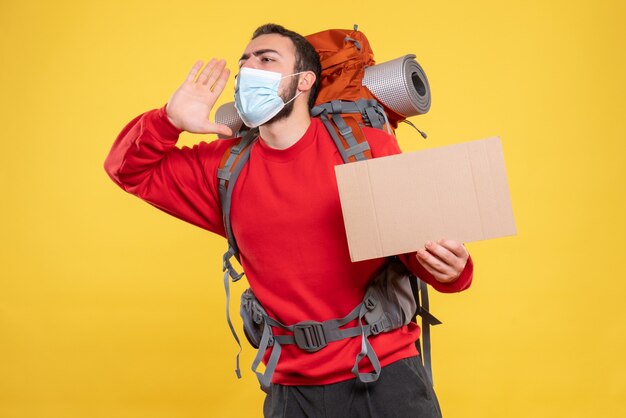 Vue de face d'un voyageur portant un masque médical avec sac à dos montrant une feuille sans écrire appelant quelqu'un sur fond jaune