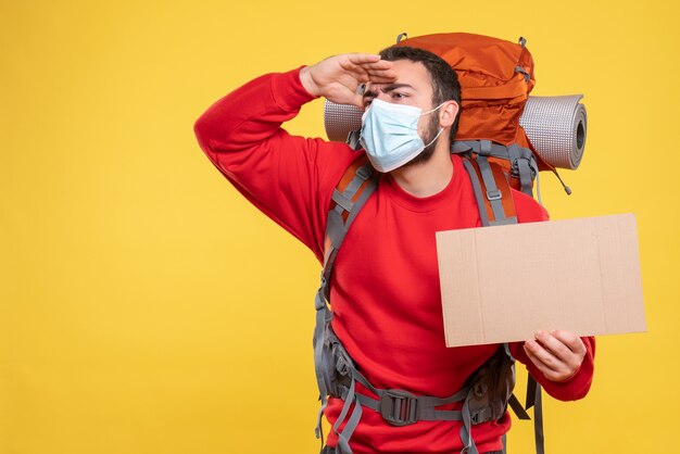 Vue de face d'un voyageur portant un masque médical avec sac à dos montrant une feuille sans écrire en regardant quelque chose avec soin sur fond jaune