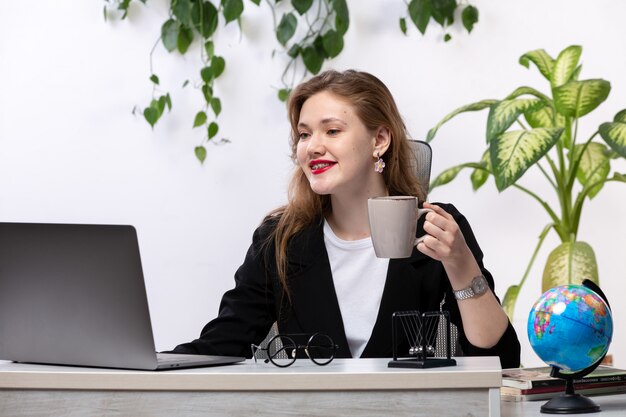 A, vue frontale, jeune, belle dame, dans, chemise blanche, et, veste noire, utilisation, elle, ordinateur portable, devant, table, sourire, tenue, tasse, à, feuilles, pendre