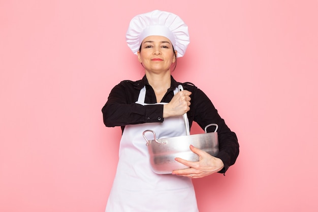 A, vue frontale, jeune femme, cuisinier, dans, chemise noire, blanc, cuisinier, cap, casquette blanche, poser, tenue, mélanger, haut, casserole argent, sourire