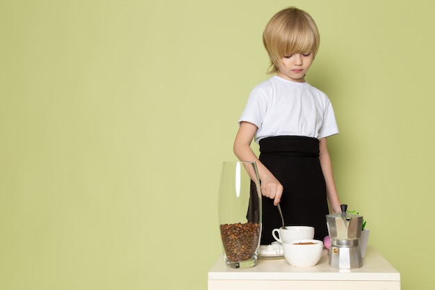 Une vue de Frotn blonde kid adorable doux mignon préparer un verre de café sur la table en t-shirt blanc sur l'espace de couleur pierre