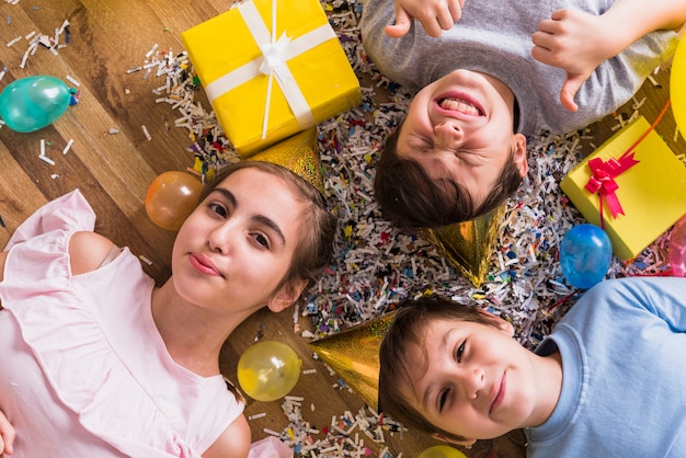 Photo gratuite vue grand angle d'amis couchés sur un plancher en bois entourés d'un cadeau; ballon et confettis