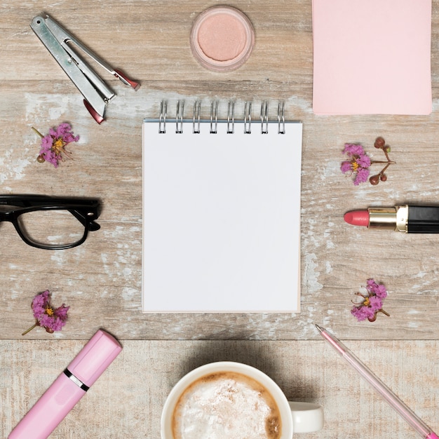 Vue grand angle de bloc-notes blanc vierge entouré de produits cosmétiques; tasse à café; fleurs et lunettes disposées sur un bureau en bois