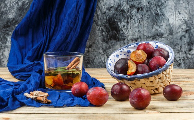 Vue grand angle boisson fermentée et cannelle sur foulard bleu avec un bol de prunes sur planche de bois et surface en marbre gris foncé. horizontal