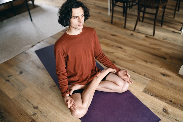 Vue grand angle du jeune homme concentré avec un corps flexible assis en posture de lotus sur un tapis, méditant avec les yeux ouverts, havinf regard attentif, se concentrant sur un objet, corps relaxant, ralentissant