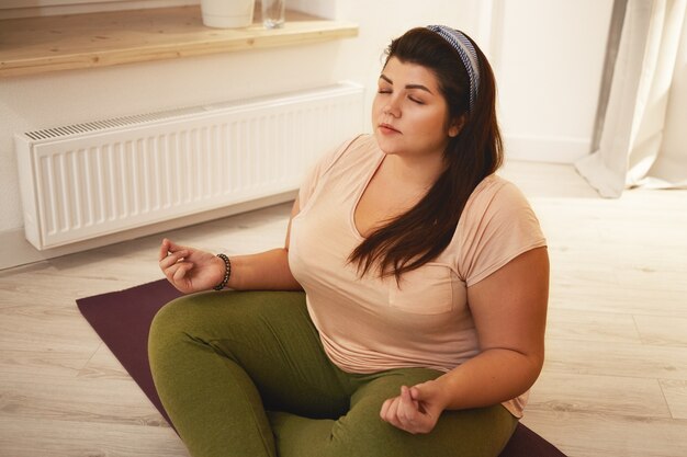 Vue grand angle de l'élégante jeune femme en surpoids potelée vêtue de jambières et t-shirt méditant avec les jambes croisées, fermant les yeux, se tenant la main en mudra, pratiquant des techniques de respiration