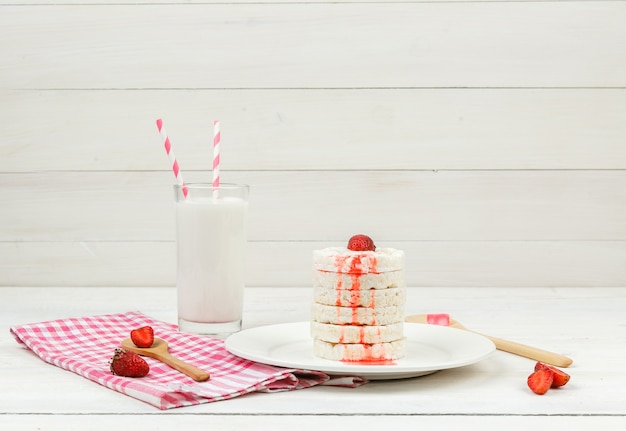 Vue grand angle des gaufrettes de riz blanc sur plaque avec des fraises, des cuillères en bois et du lait sur la surface de la planche de bois blanc.