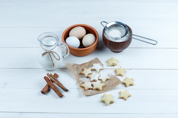 Vue grand angle des oeufs dans un bol avec des biscuits, des bâtons de cannelle, du lait, du sucre en poudre sur fond de bois.