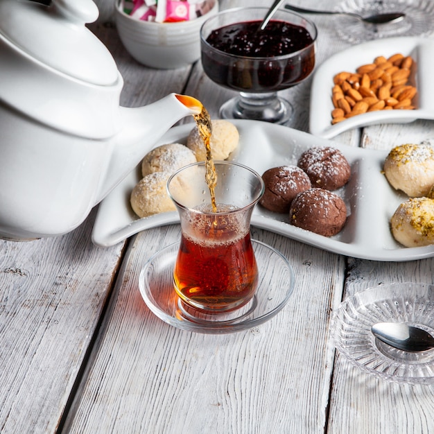 Vue grand angle quelqu'un versant du thé avec des desserts, des noix, de la confiture de fruits sur fond de bois blanc.