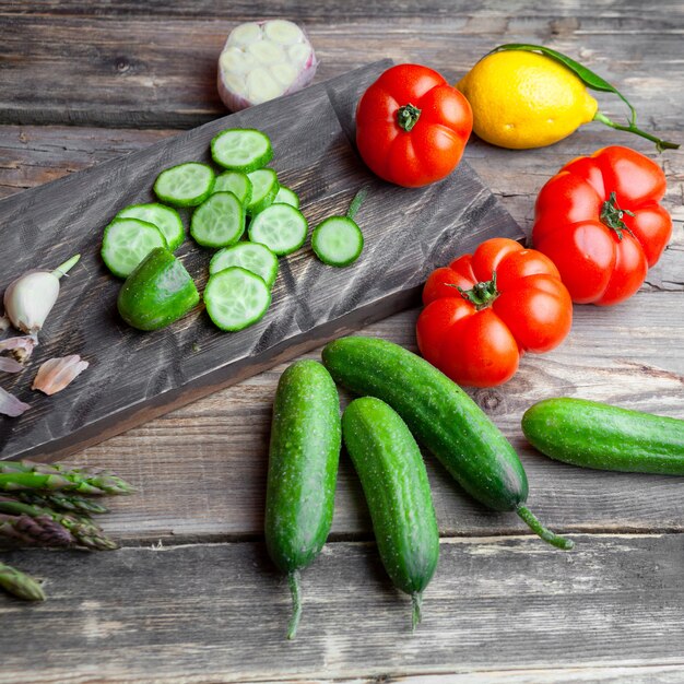 Vue grand angle en tranches de concombre en planche à découper avec de l'ail, des tomates, de la laitue, du citron sur fond de bois foncé.