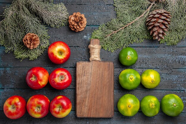 Vue en gros plan de dessus de pommes limes six planches à découper de pommes jaune-rougeâtre et six limes sur une surface grise à côté des branches et des cônes d'épinette