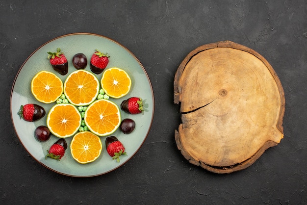 Vue en gros plan des fruits et de l'orange hachée au chocolat avec des fraises enrobées de chocolat et des bonbons verts à côté d'une planche à découper sur une surface sombre