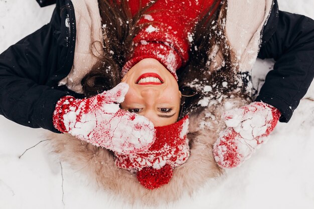 Vue d'en haut sur jeune femme heureuse souriante assez candide dans des mitaines rouges et bonnet tricoté portant un manteau noir couché dans la neige dans le parc, des vêtements chauds, s'amuser